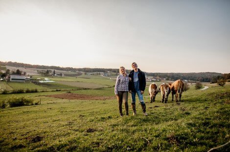 Two people standing on a hill in a field with three brown and white cows behind them
