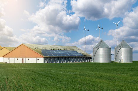 Dairy farm with a barn that has solar panels on the roof, two silos, and three wind turbines in the background