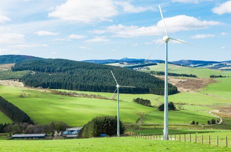Two wind turbines on a fence line in a grass field, with farm buildings, hills, and a forest in the background